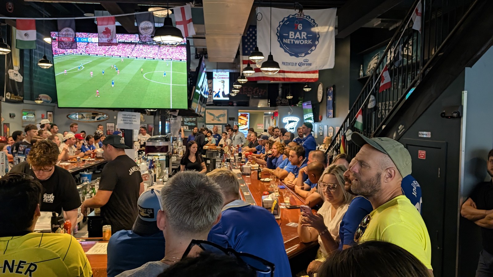 A wide view down the bar at Cavanaugh's with every seat along it taken, flags and banners hanging from the ceiling and the match on the overhead screens.