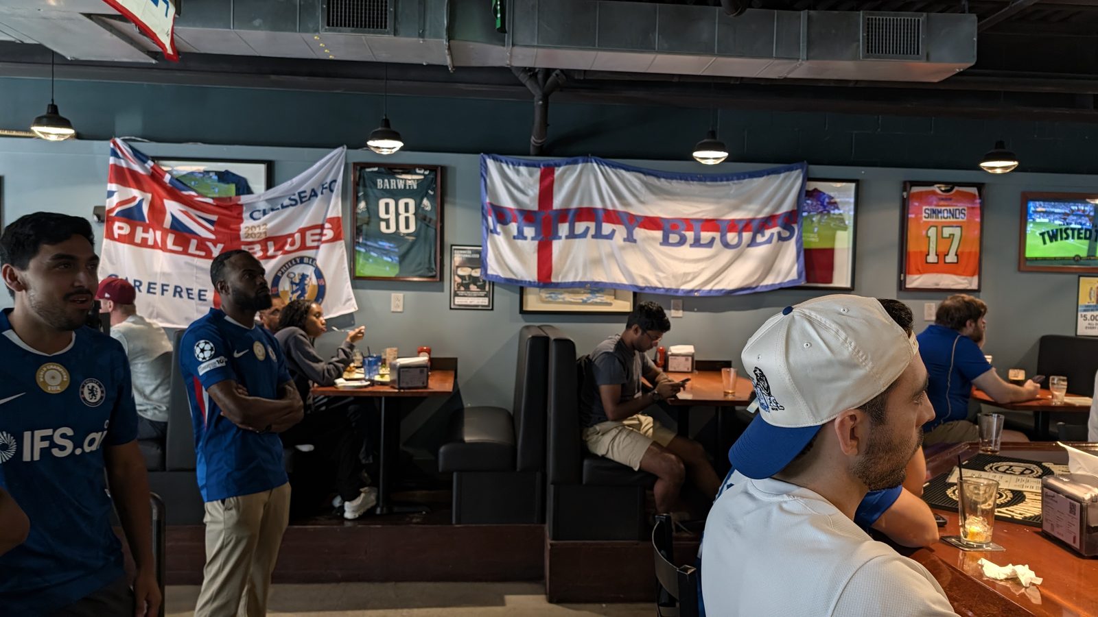 Two Philly Blues flags hung on the wall between framed Philadelphia sports jerseys, with supporters watching the match nearby.