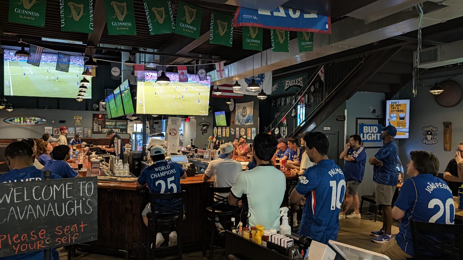 The bar at Cavanaugh's Rittenhouse packed with supporters in blue Chelsea shirts watching the match on the screens above the bar. A chalkboard reads Welcome to Cavanaugh's, please seat yourself.