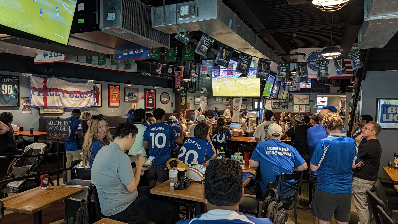 A wide view of the room with tables and booths full, the Philly Blues flag hanging on the wall, and the match playing on the overhead screens.